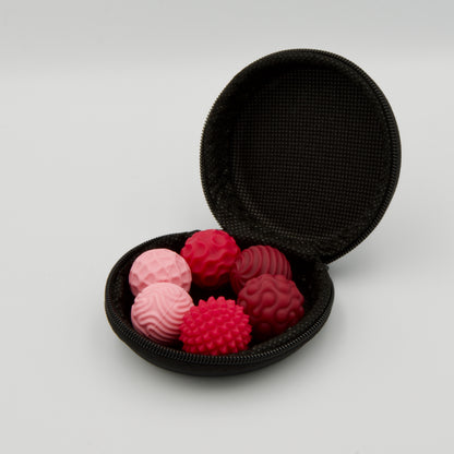 Geöffnetes, schwarzes Rund-Etui in schräger Seitenansicht, gefüllt mit sechs roten und rosa Haptik-Bällen mit unterschiedlichen Oberflächenstrukturen