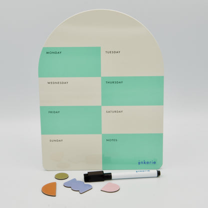 Bogenförmige magnetische Wochenplaner-Tafel von inkerie in Mintgrün und Beige mit Stift und vier bunten Formmagneten davor.