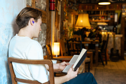 Junger Mann sitzt lesend mit einem Buch in einem Café und trägt einen blauen Gehörschutz-Stöpsel im Ohr.