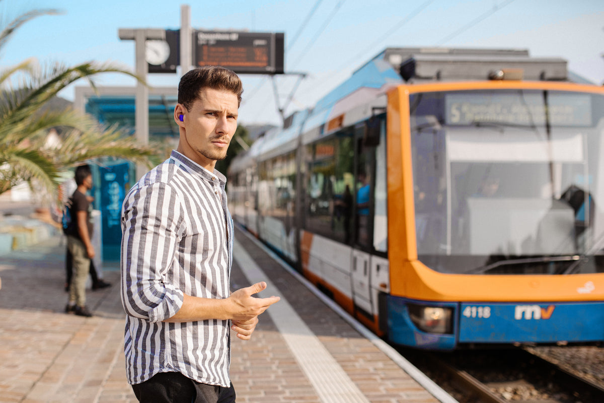 Mann im gestreiften Hemd an einer Bahn-Haltestelle mit einem blauen Gehörschutz-Stöpsel im Ohr